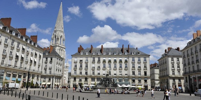 Place publique bordée d'immeubles historiques européens, une flèche d'église élancée, une fontaine et des passants sous un ciel bleu partiellement nuageux